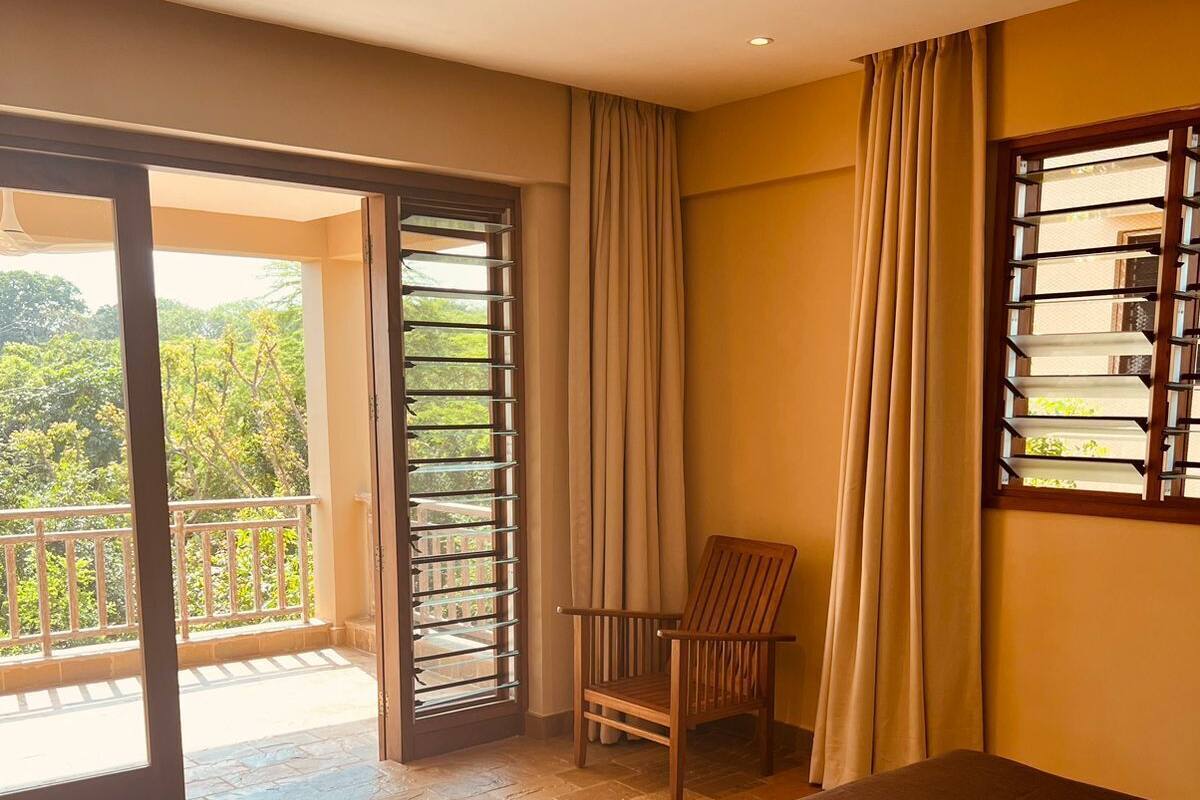 A hotel room with a bed, a chair, a balcony with a view of greenery, and a ceiling fan.