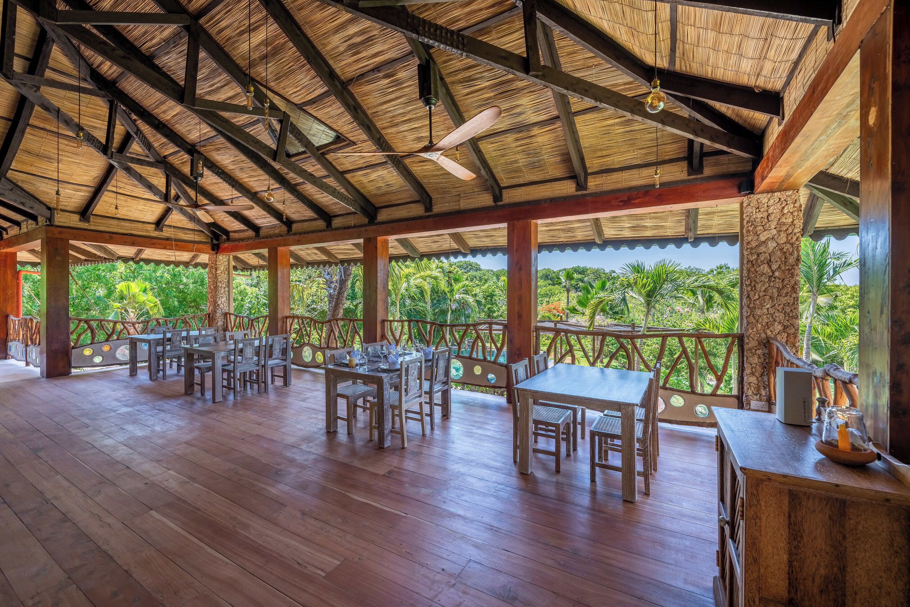 A covered outdoor dining area with wooden tables and chairs, a ceiling fan, and a view of lush greenery.