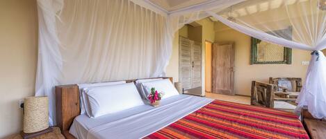 A bedroom with a bed, white curtains, a wooden headboard, a bedside table with a lamp, and a colorful striped bedspread.