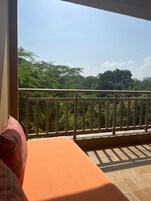 A balcony with a wooden railing, a cushioned seating area, and a view of lush greenery.