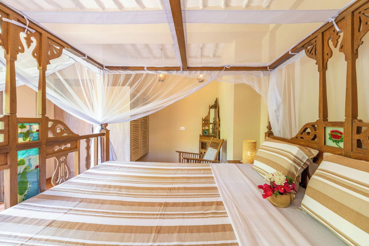 A bed with striped bedding, a wooden headboard, and a canopy. There is a chair and a mirror in the background.