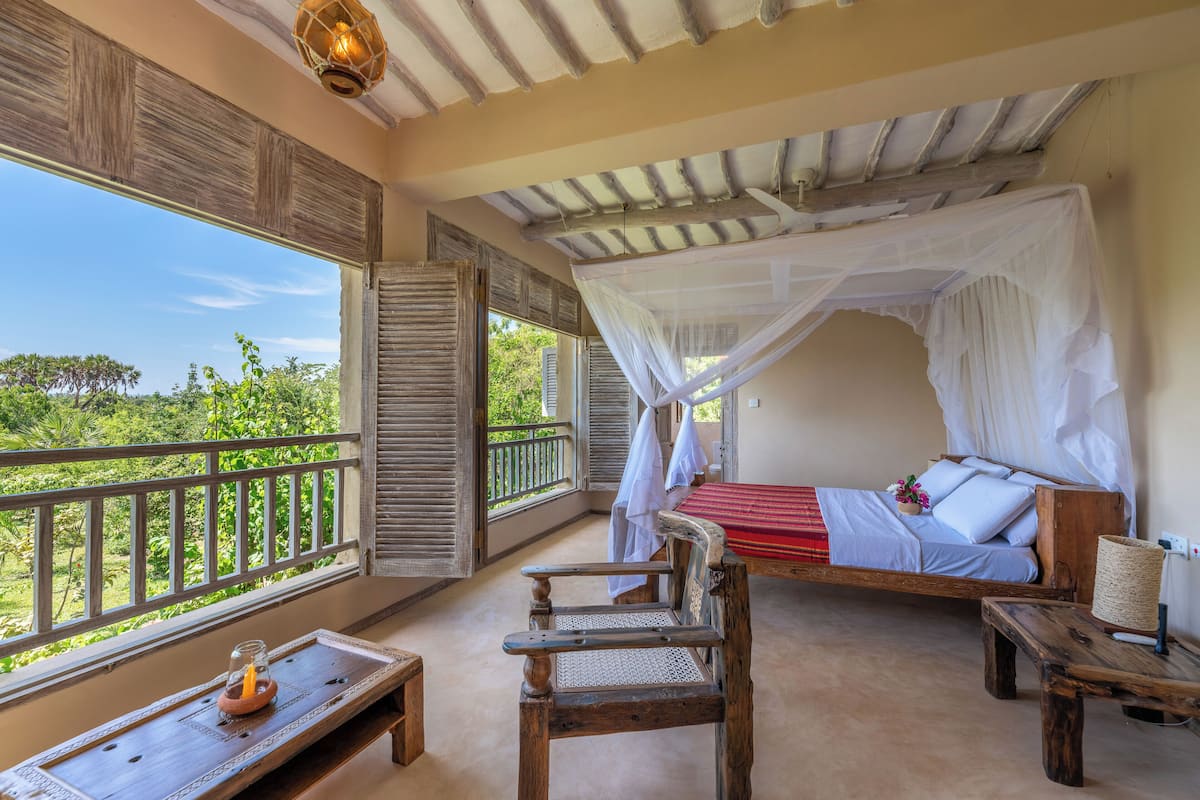 A room with a bed under a canopy, a wooden chair, a small table, and a view of greenery outside.