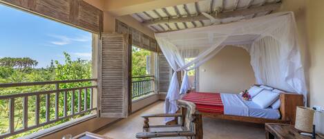Habitación con cama bajo un dosel, una silla de madera, una mesita y vistas al exterior con vegetación.