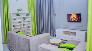 Un salon comprenant un canapé, une petite table ronde, un téléviseur fixé au mur, un meuble blanc et une étagère avec des plantes.