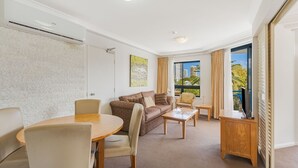 Et hotelværelse med sofa, stole, et spisebord, et fjernsyn og et stort vindue med udsigt til bygninger og palmer.