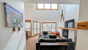 A modern living room with a sectional sofa, a wooden coffee table, and a fireplace. There are large windows providing natural light and a view of the outdoors. - Large Bright Condo w/ Hot Tub, Pool & Shuttle (Keystone)