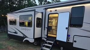 Una caravana con una rampa que lleva a la puerta, un interior visible con una cama, y una ventana que muestra una vista al atardecer.