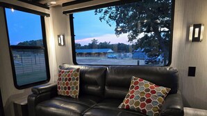 Un sofá de cuero negro con dos cojines estampados, una ventana con vista a un estacionamiento y árboles, y luminarias fijadas a la pared.