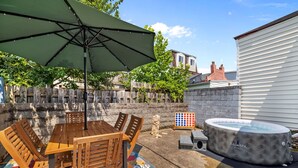 Une terrasse avec une table et des chaises en bois, un parasol vert, un jacuzzi et un tapis coloré.