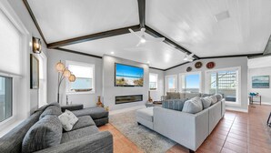 A spacious living room with a fireplace, large grey sofas, and a high ceiling with exposed beams.