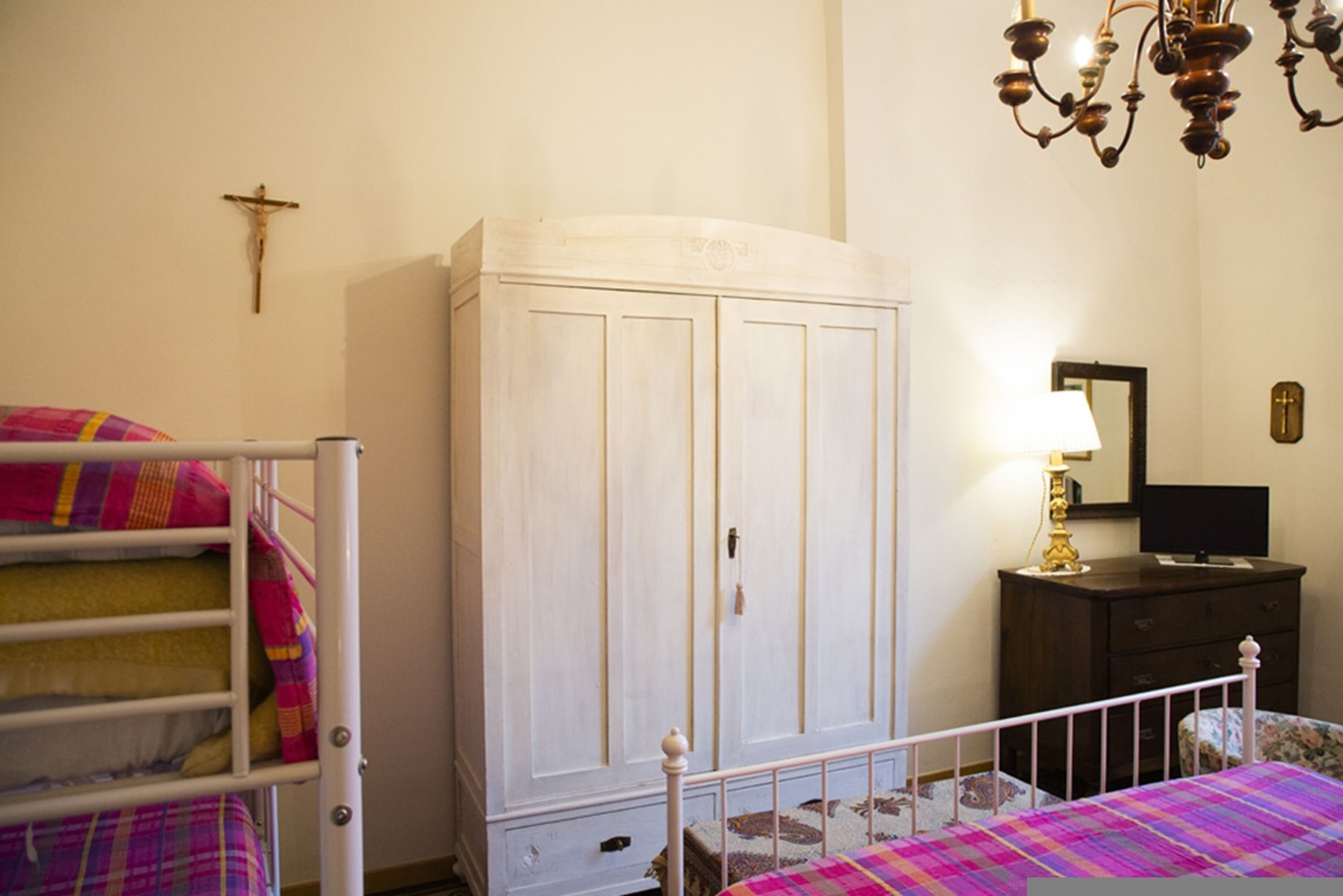 Une chambre avec des lits superposés, une armoire, un bureau avec une lampe et une croix accrochée au mur.