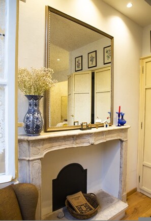 A fireplace with a mirror above it, a vase with flowers, and a basket with a newspaper on the mantel.