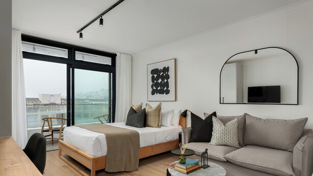A modern hotel room with a large bed, a sofa, a small table, and a view of the city through large windows. - The Sage - Elegant Studio in Vibrant Sea Point (Cape Town)