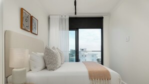A modern hotel room with a large bed, a bedside table with a lamp, and a view of the city through a large window. - The Sage l Designer One Bedroom l Ocean Views (Cape Town)