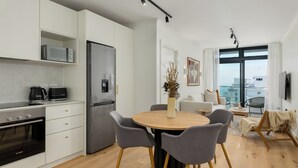 A modern kitchen with white cabinetry, a built-in microwave, a stainless steel refrigerator, and a Bosch oven. There is a round wooden table with four grey chairs and a vase with dried plants. The room has a large window with a view of the city. - The Sage l Designer One Bedroom l Ocean Views (Cape Town)