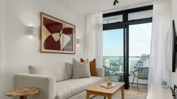 A modern living room with a sofa, a small wooden table, and a balcony with a view of the city. - The Sage l Designer One Bedroom l Ocean Views (Cape Town)