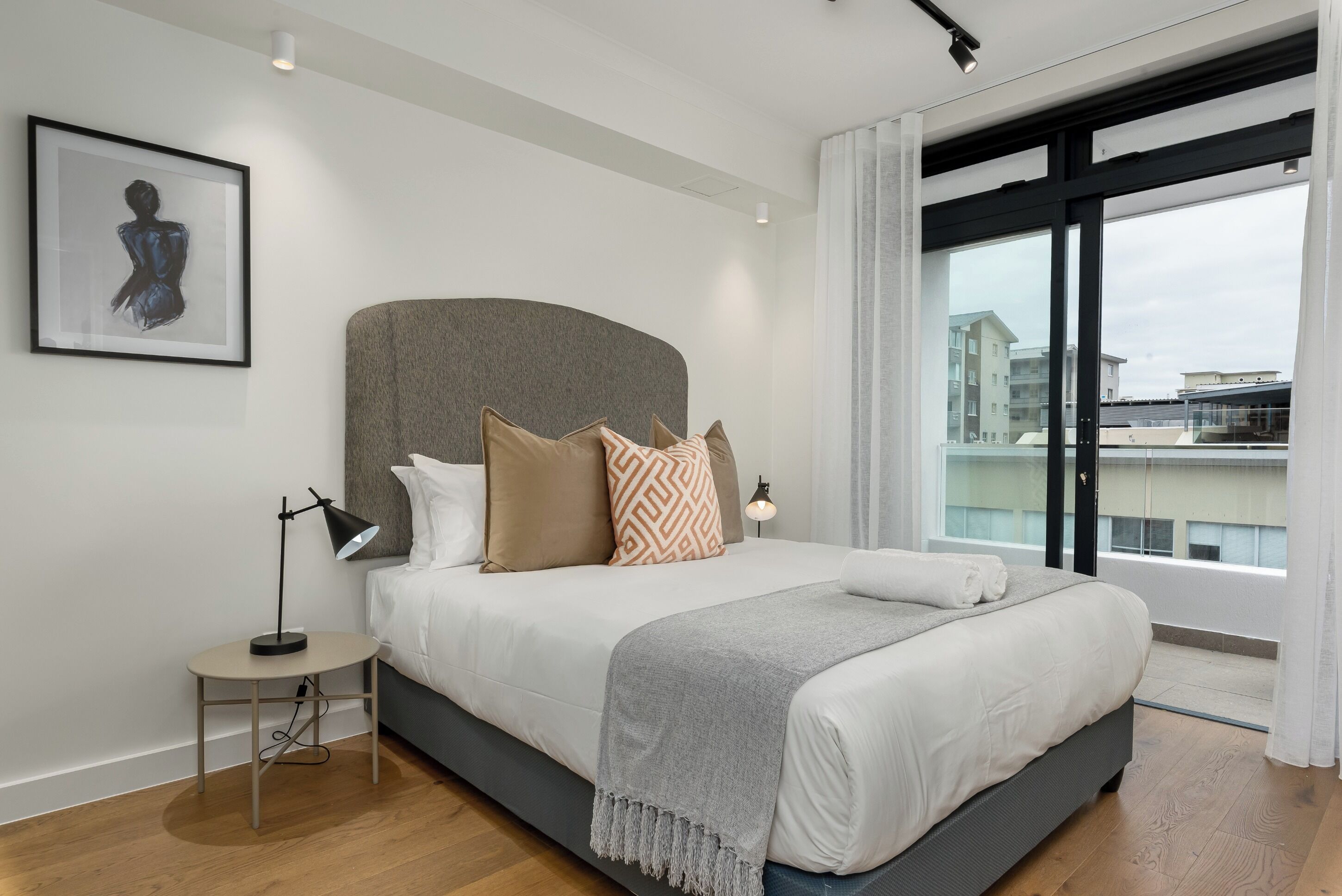 A modern bedroom with a large bed, a bedside table with a lamp, a framed artwork on the wall, and a view of buildings through the window.