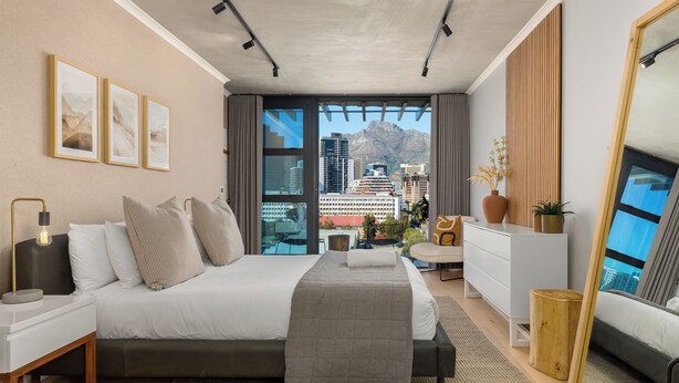 A modern bedroom with a large bed, a nightstand, a dresser, and a view of a cityscape through large windows.