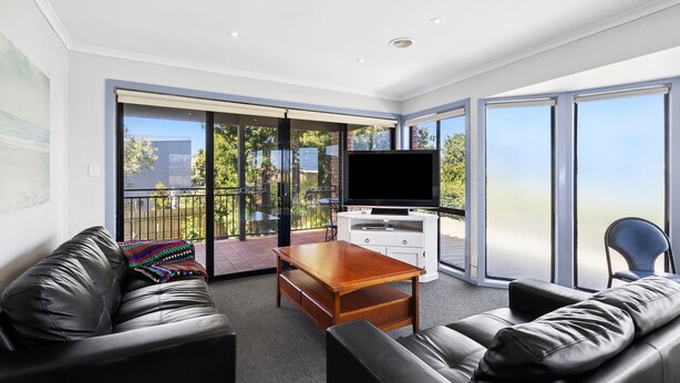 Ein modernes Wohnzimmer mit einem Flachbildfernseher, Ledersofas, einem Holztisch und großen Schiebegläsern, die auf einen Balkon mit Blick führen.