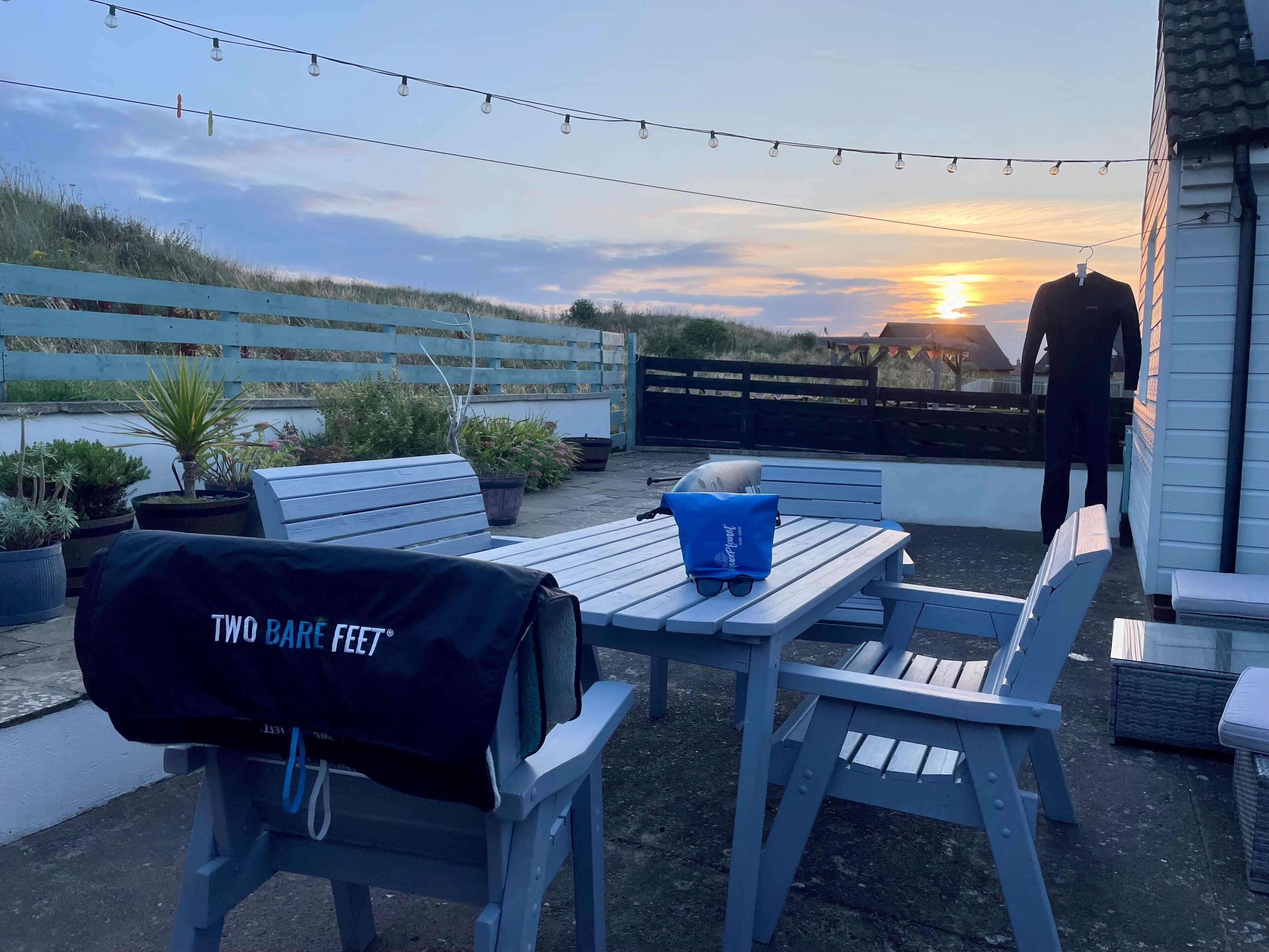 An outdoor patio with a table and chairs, a barbecue grill, string lights, and a view of the sunset.