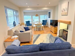 A modern living room with a sofa, a dining table with chairs, a fireplace, and a television. - Marram Cottage - Cosy, well equipped, family, beach house (Chathill)