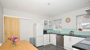 A modern kitchen with white cabinets, a wooden table, and a clock on the wall. - Marram Cottage - Cosy, well equipped, family, beach house (Chathill)