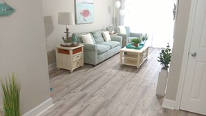 A living room with a light-colored wooden floor, a green sofa, a white side table, and a small white cabinet. There is a painting on the wall and a door to the right. - Charming BEACHFRONT condo in Galveston, 2 POOLS open year round, FITNESS CENTER (Galveston)