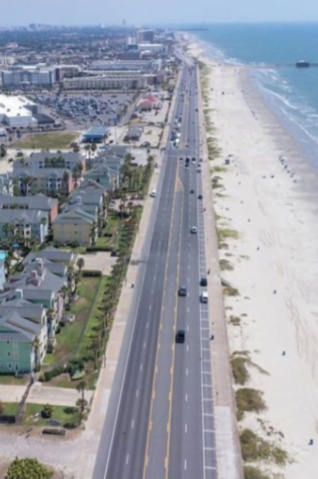 A coastal road with a beach on one side and residential buildings on the other.