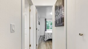 Ein schmaler Flur mit weißen Wänden und Holzböden, der zu einem Schlafzimmer mit Bett und Fenster führt.