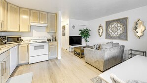Eine moderne Küche mit weißen Geräten, ein Essbereich mit weißem Tisch und Stühlen sowie ein Wohnzimmer mit grauem Sofa und Fernseher.