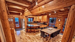 A log cabin interior with a kitchen, dining area, and a large window overlooking a forested area.