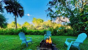 A fire pit with three Adirondack chairs arranged around it in a grassy outdoor area. - Charming cottage on the ORANGE RIVER in Fort Myers Fl. Pool ,Kayaks, AC, WiFi (Fort Myers)
