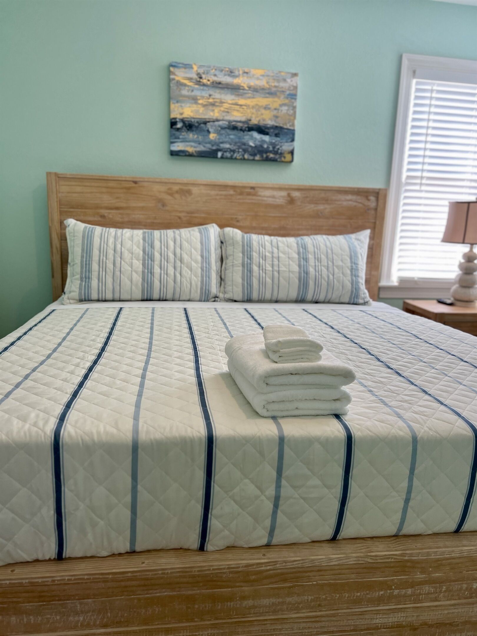 A bed with a quilted bedspread and pillows, a painting on the wall, and a window with blinds.