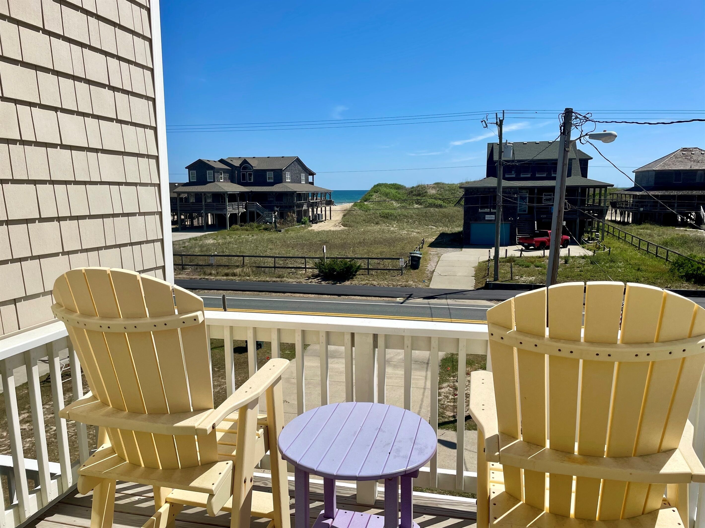 Een balkon met twee gele Adirondack-stoelen en een kleine paarse tafel, met uitzicht op een strand met huizen en een weg.