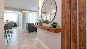 A modern living room with a wooden shelf holding plants, a large mirror, and a dining area with chairs and a table. - Stylish 2-br Apt Sleeps 7 Near Disney Magic (Kissimmee)