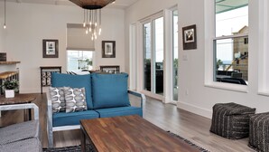 A spacious living room with a blue sofa, wooden coffee table, and a large window offering a view of the outdoors. - Rejuvenation:Brand New, Beach Views, Custom Furniture, Hot Tub + MIL Suite (St. Joe Beach)