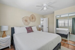 Habitación de hotel con una cama grande, dos mesitas de noche con lámparas, un ventilador de techo, un espejo amplio y vistas al exterior a través de una ventana.