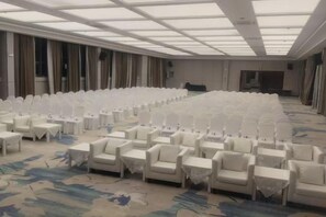 Une grande salle de conférence avec de longues rangées de chaises et de tables blanches, un sol recouvert de moquette et un plafond haut doté de lucarnes.