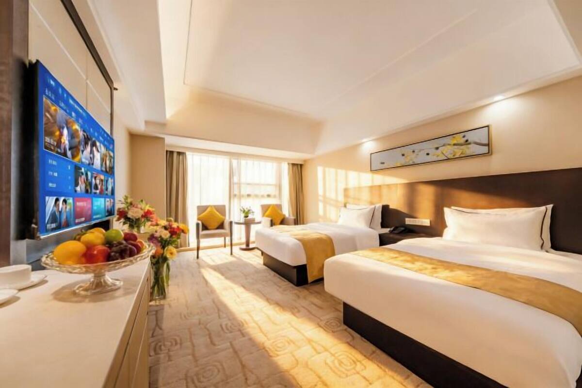 Habitación de hotel con dos camas, televisión, escritorio con frutero y un jarrón con flores.