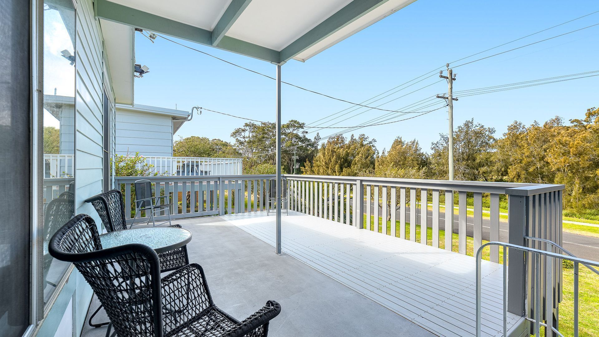 A covered balcony with a table and chairs, a railing, and a view of the street and trees.