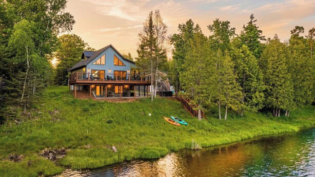 A lakeside cabin with a deck, surrounded by trees and a river.