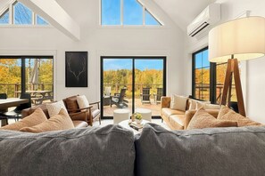 A spacious living room with a large sofa, a coffee table, and a dining area with a table and chairs. There are large windows offering a view of the outdoors.