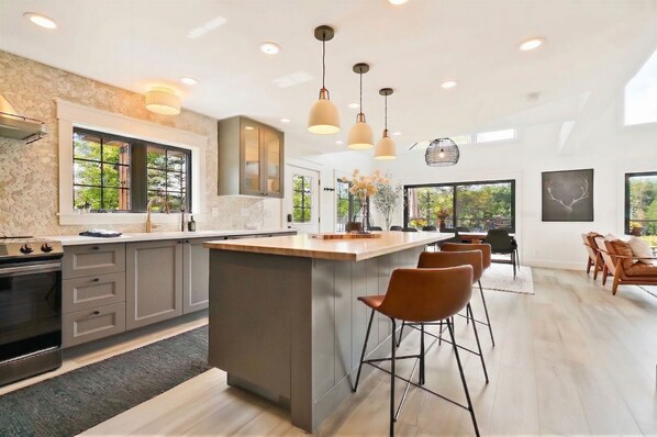 A modern kitchen with a central island, bar stools, and large windows. - Luxury Riverfront Lodge · Hot Tub · Sleeps 12 (Cornell)