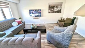 A modern living room with a grey sofa, patterned armchair, and a coffee table with decorative items. A flat-screen TV is mounted on the wall, and a cityscape painting is visible. - Niagara Getaway 
4bdrm, 4bths. 18 min to Falls.
'Isla Paraiso Verde' (Thorold)