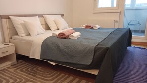 Un lit bien fait, recouvert d’une couverture grise, avec des oreillers blancs et des serviettes enroulées. On aperçoit une table de chevet avec une lampe et un petit tiroir. Un tapis à motifs recouvre le sol, et une fenêtre avec des stores est visible au fond.
