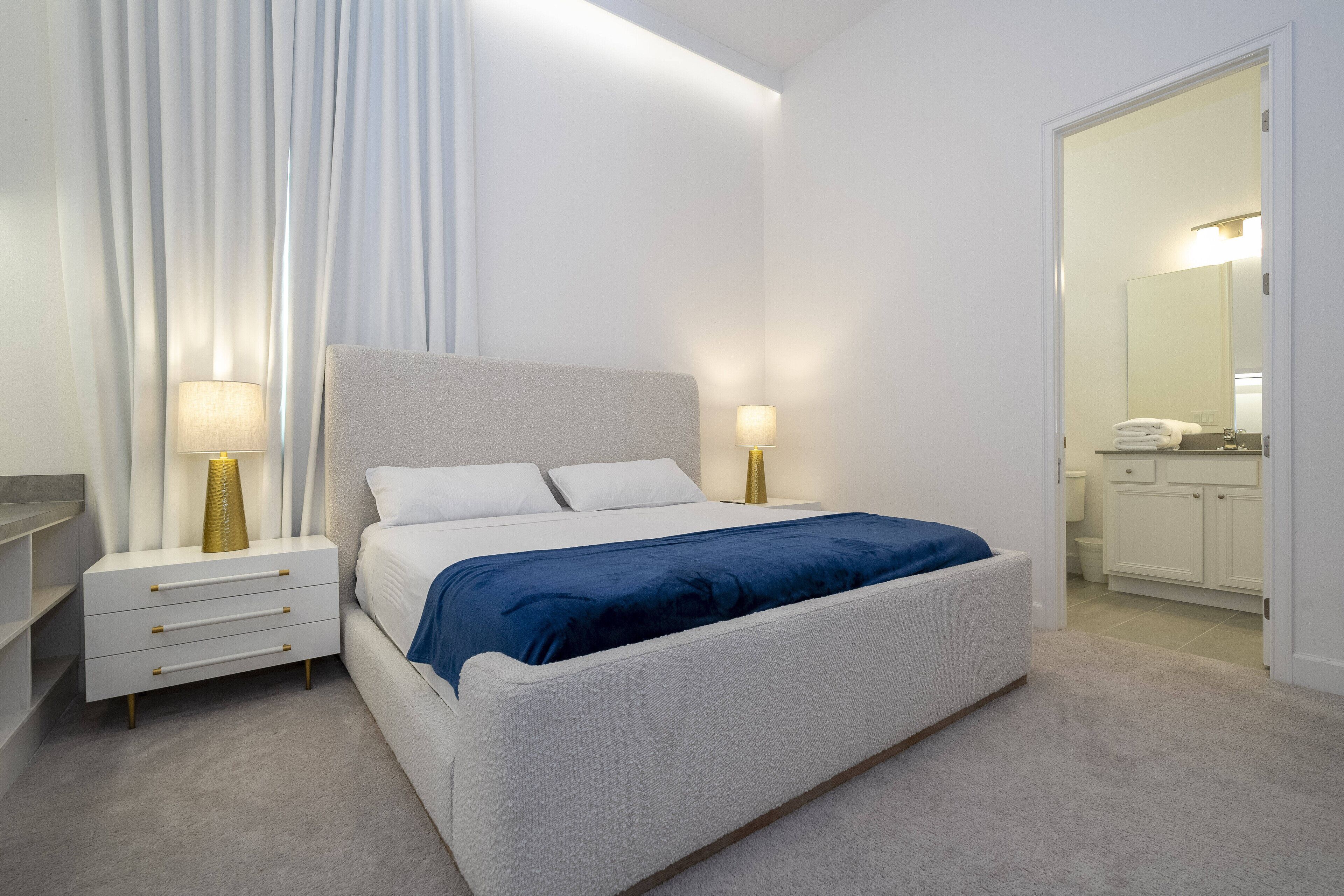 Un dormitorio moderno con cama, mesitas de noche y un baño visible a través de una puerta abierta.