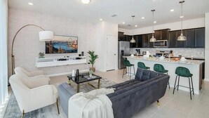 Un salon moderne avec un canapé, des fauteuils, une table basse et un téléviseur. La cuisine est dotée de meubles de couleur sombre, d’un four à micro-ondes et d’un évier.