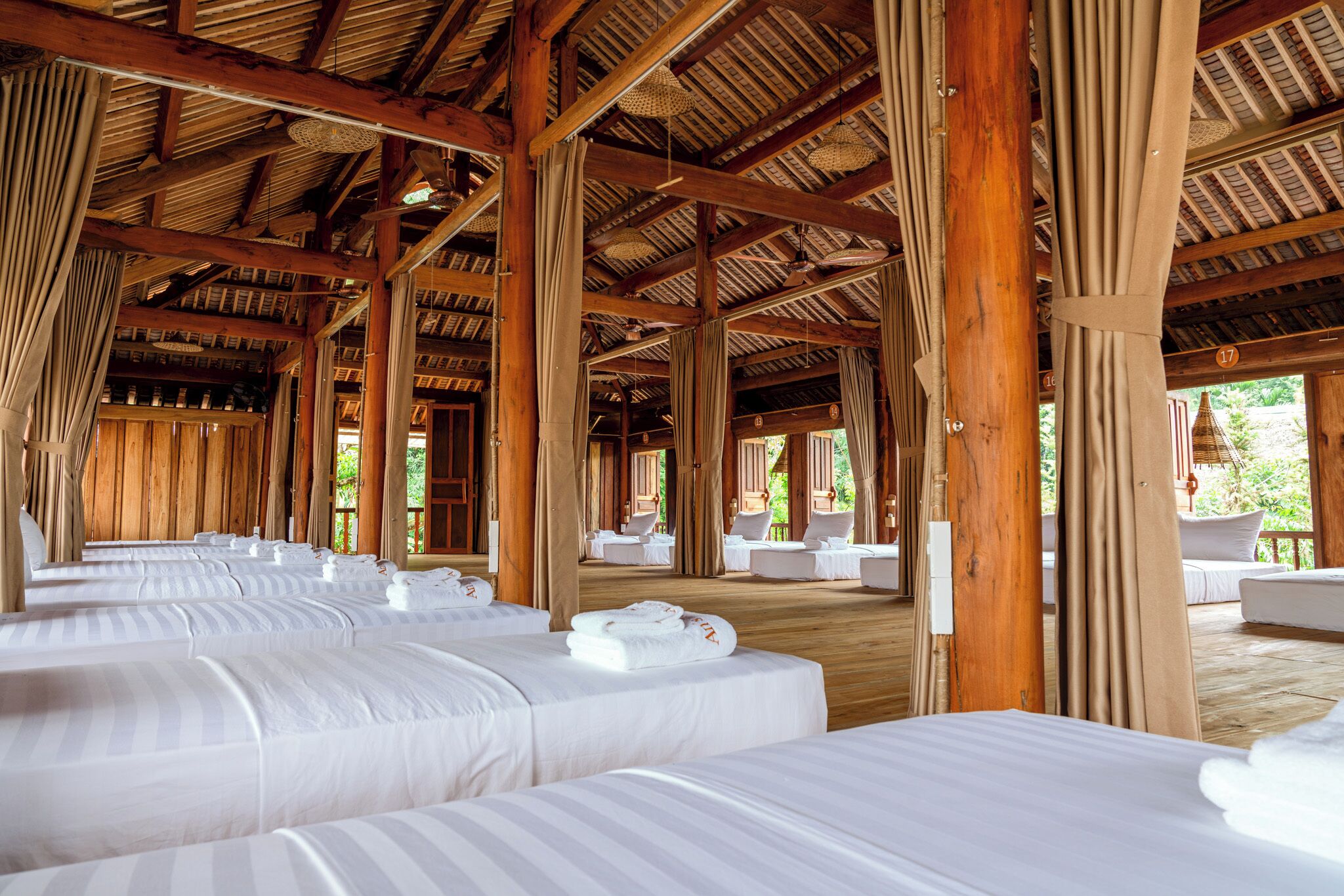 A large, open room with multiple beds arranged in rows, wooden beams on the ceiling, and a view of the outdoors.