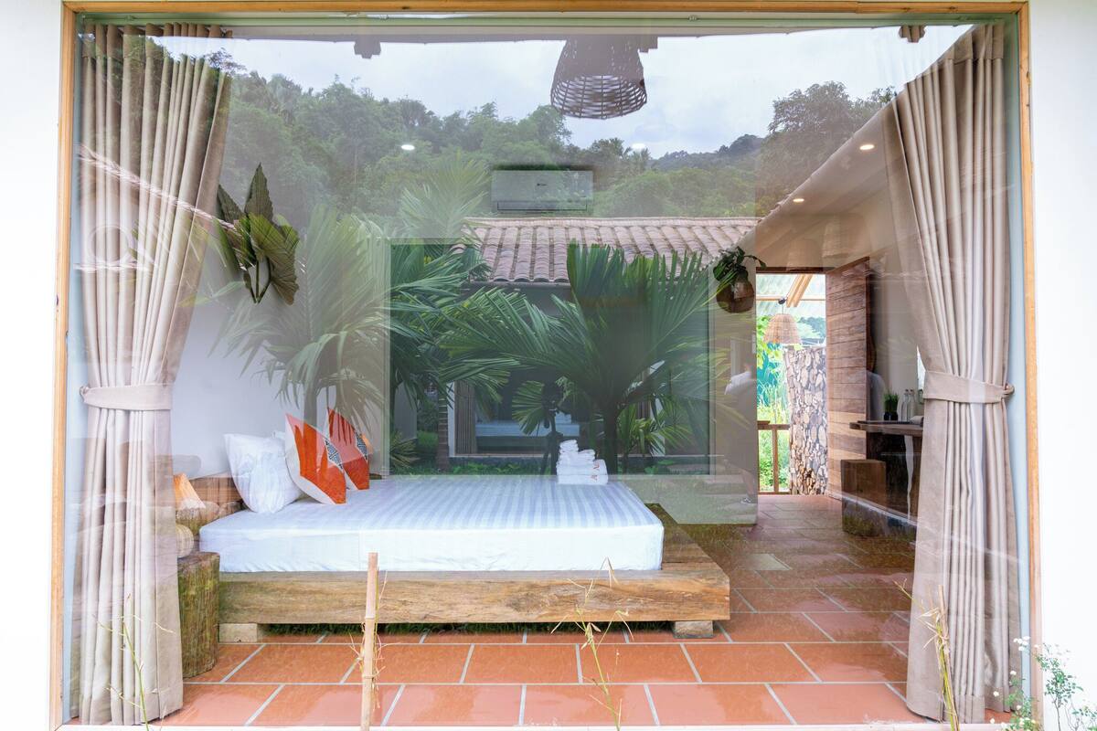 Ein Schlafzimmer mit Bett, Vorhängen, einer Pendelleuchte und Blick auf Grünflächen.