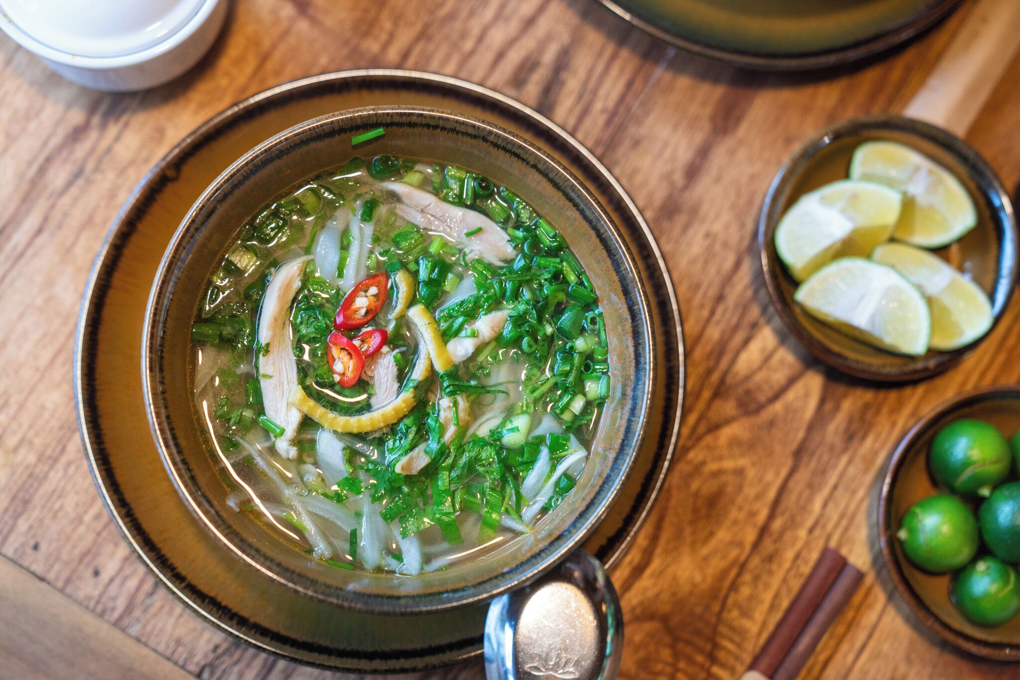 Un tazón de sopa con varios ingredientes, acompañado de un tazón pequeño de rodajas de lima y un tazón de limas verdes.